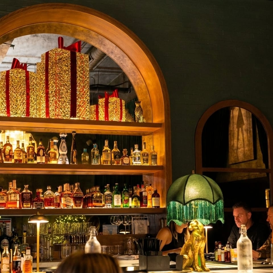 Warmly lit restaurant bar with liquor shelves, decorative gift boxes, vintage lamps, and patrons seated at the counter.