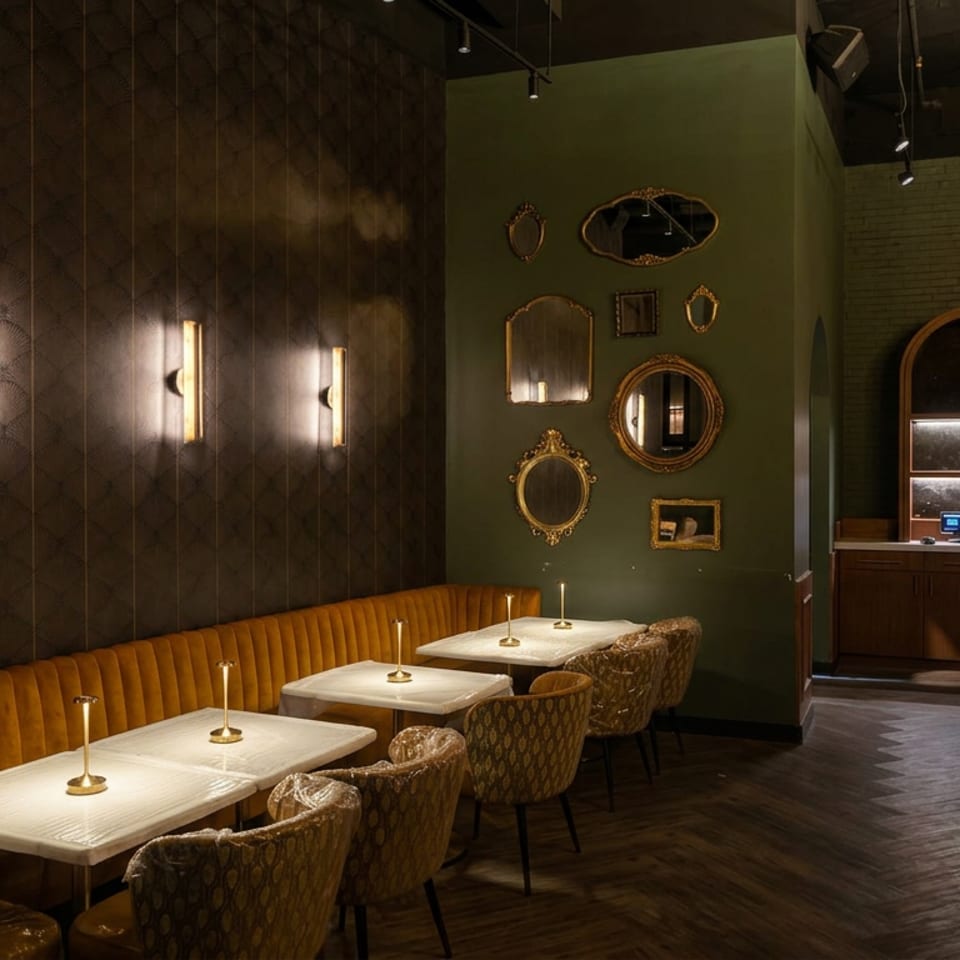 Elegant restaurant interior with mustard velvet banquette seating, marble tables, brass candles, decorative mirrors, and a well-stocked bar.