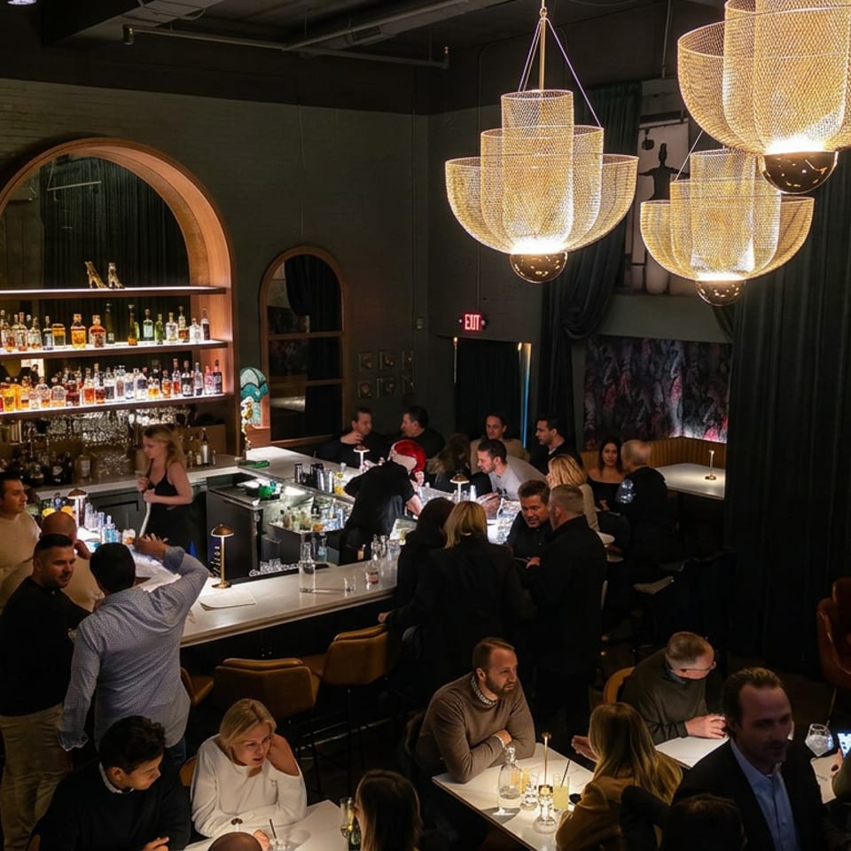 Bustling upscale bar interior with glowing pendant lights, liquor shelves, bartenders, and patrons seated at the counter and tables.