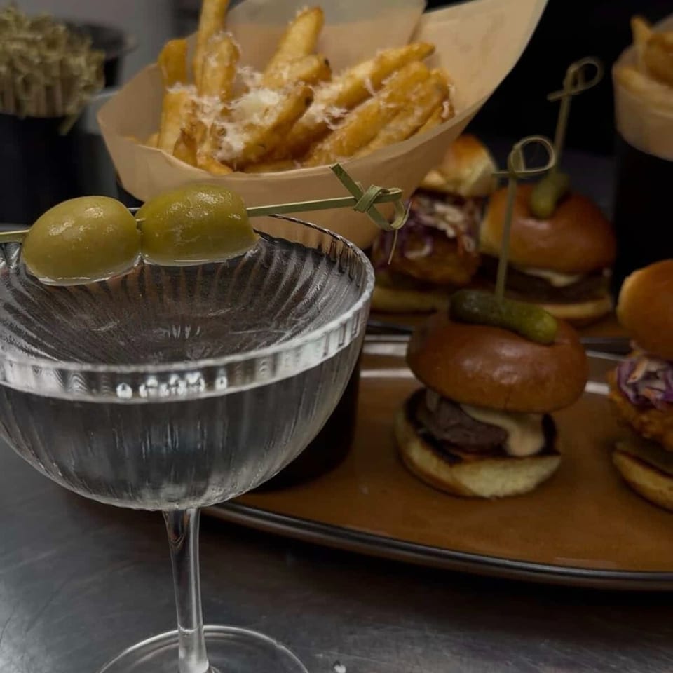 Martini glass garnished with green olives beside loaded fries and mini burgers on a restaurant serving tray.