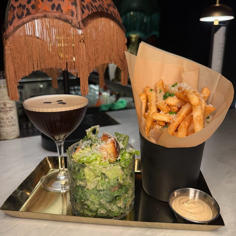 Restaurant tray featuring crispy fries with dipping sauce, Caesar salad, and a dark cocktail beneath a fringed leopard-print lamp.