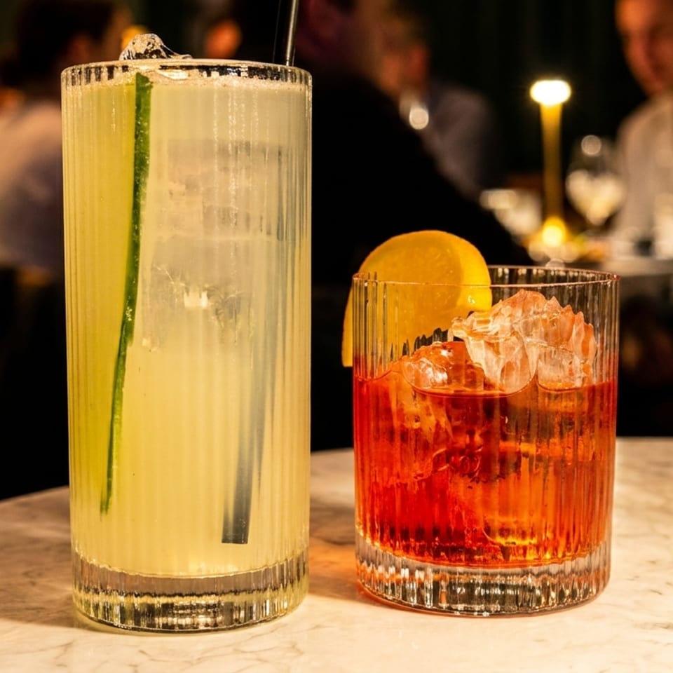 Two cocktails on a marble table: a tall pale drink with cucumber garnish and a red-orange drink with ice and orange slice.