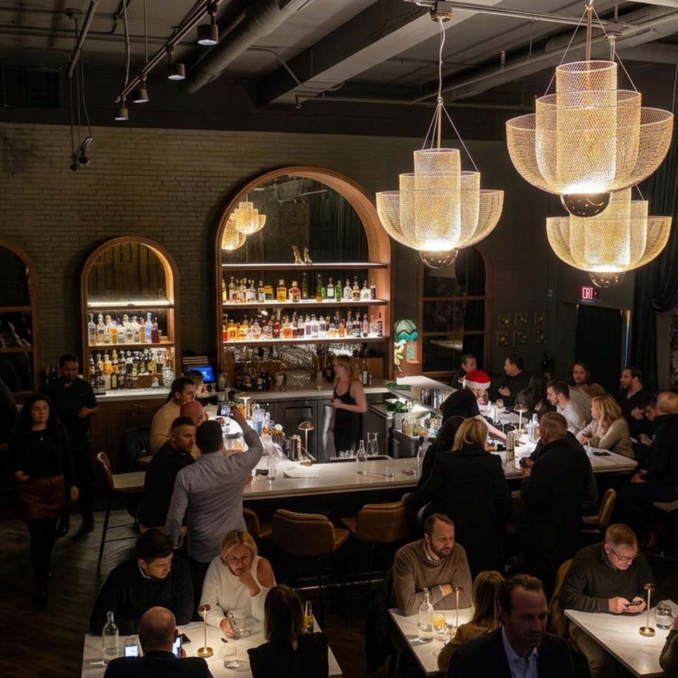 Bustling upscale restaurant with diners, a central bar stocked with bottles, exposed brick walls, and glowing tiered chandeliers.