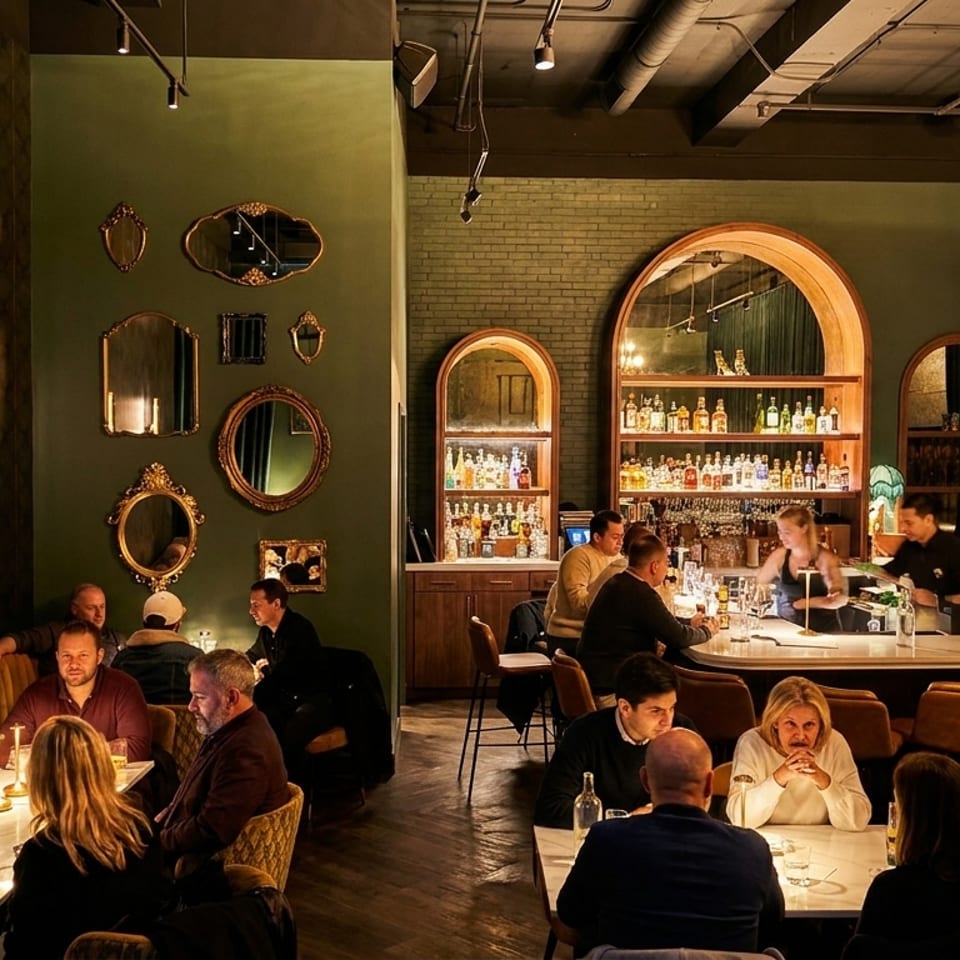 Warmly lit upscale restaurant interior with diners, a long bar, arched liquor shelves, mirrors, and intimate dining tables.