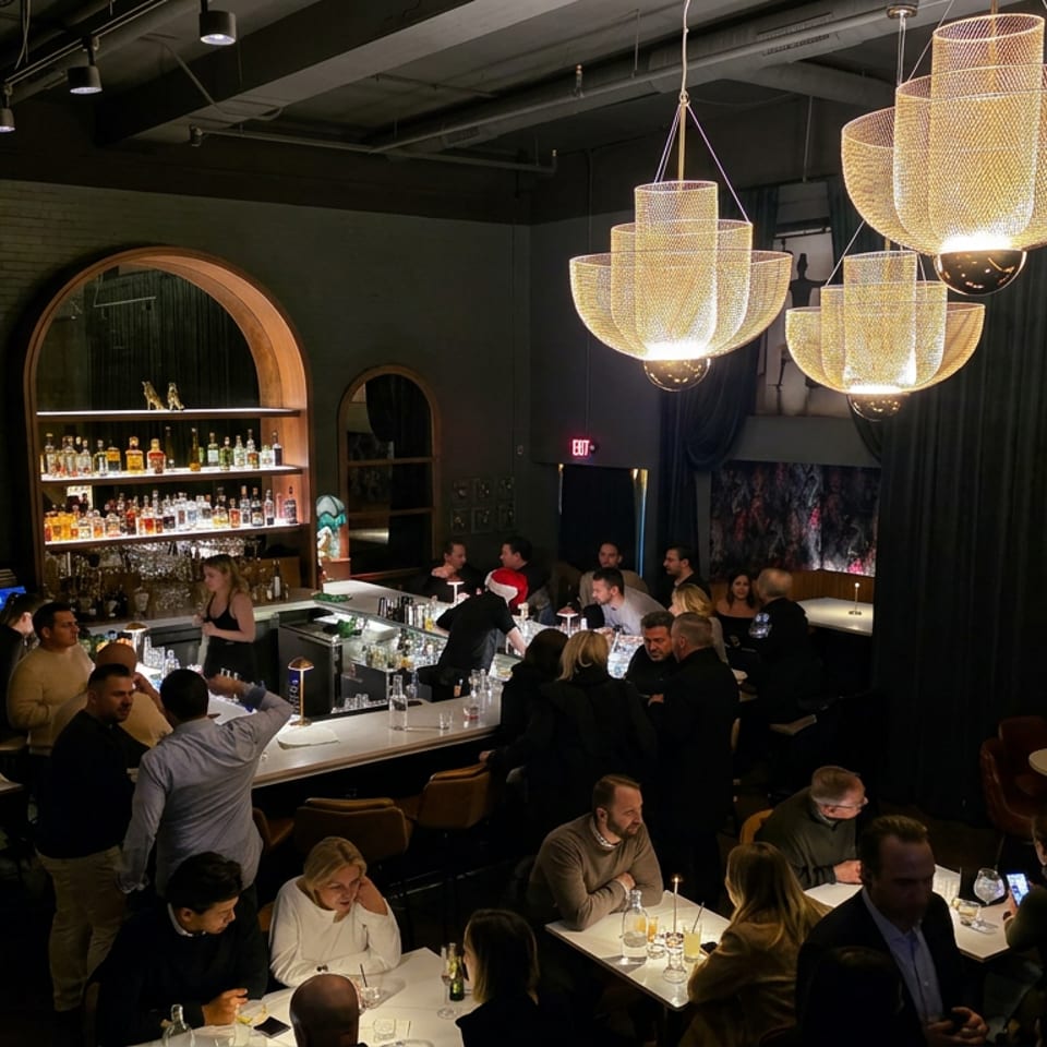 Busy upscale restaurant and bar interior with glowing chandeliers, arched liquor shelves, and diners seated at tables and the counter.