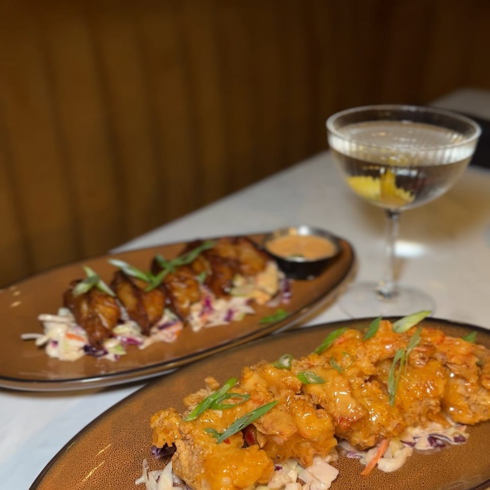 Two plates of glazed crispy chicken served over creamy slaw, garnished with green onions, alongside a cocktail in a dimly lit restaurant.