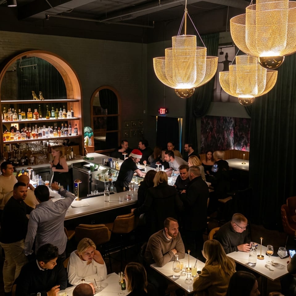 Busy upscale restaurant bar with patrons dining, illuminated sculptural chandeliers, arched liquor shelves, and a bartender serving guests.