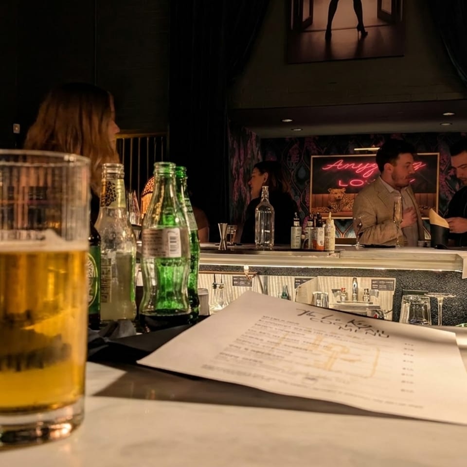 Warmly lit restaurant bar with a beer, bottled drinks, menu, patrons, bartenders, and decorative fringe lamps.
